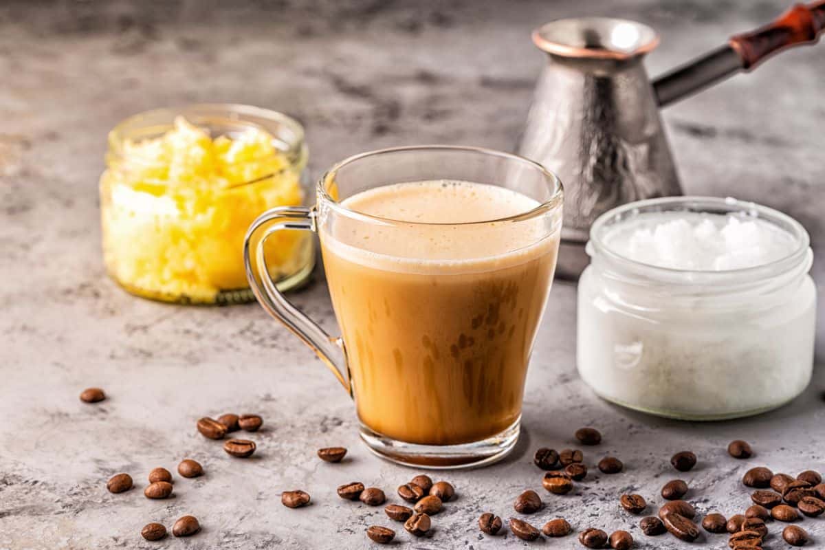 A clear glass mug of light brown frothy bulletproof coffee next to a tub of grass-fed butter and a jar of white coconut milk. Whole coffee beans surround the scene for decoration.
