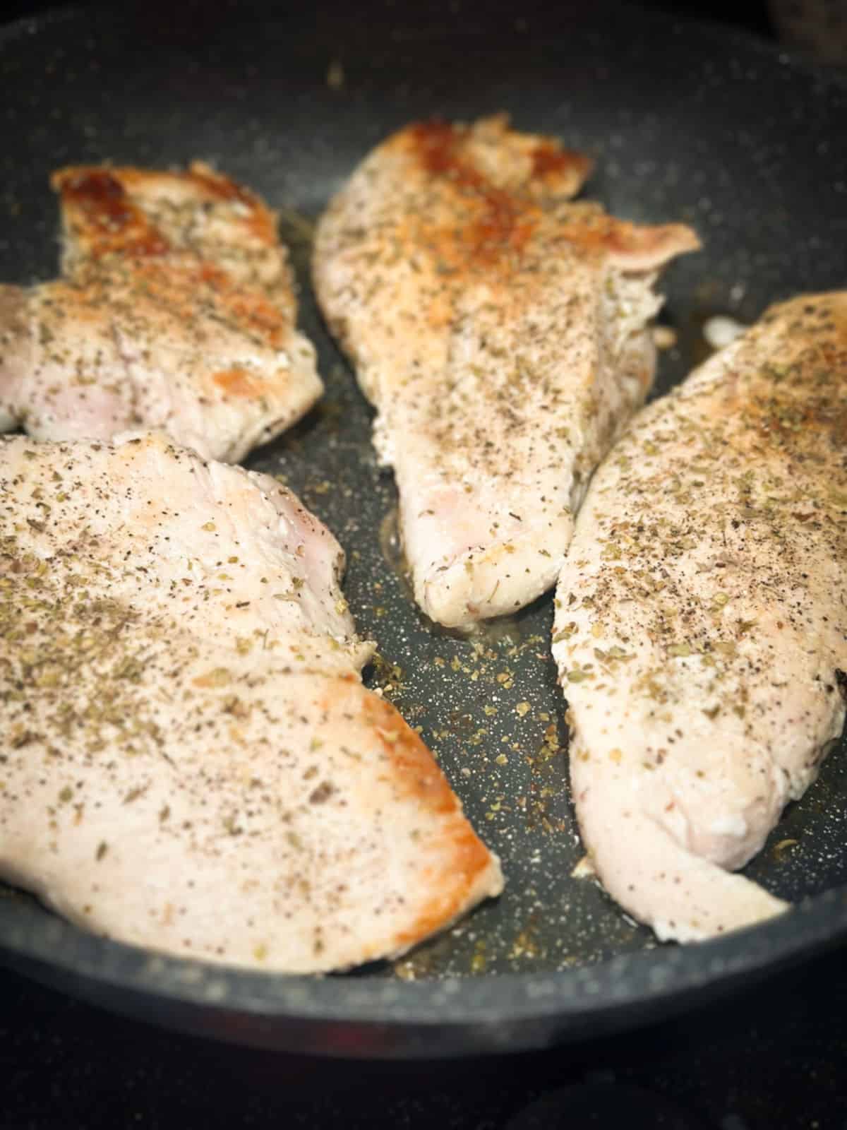 A closeup photo of chicken breast cooking in a skillet seasoned with italian seasoning salt and pepper