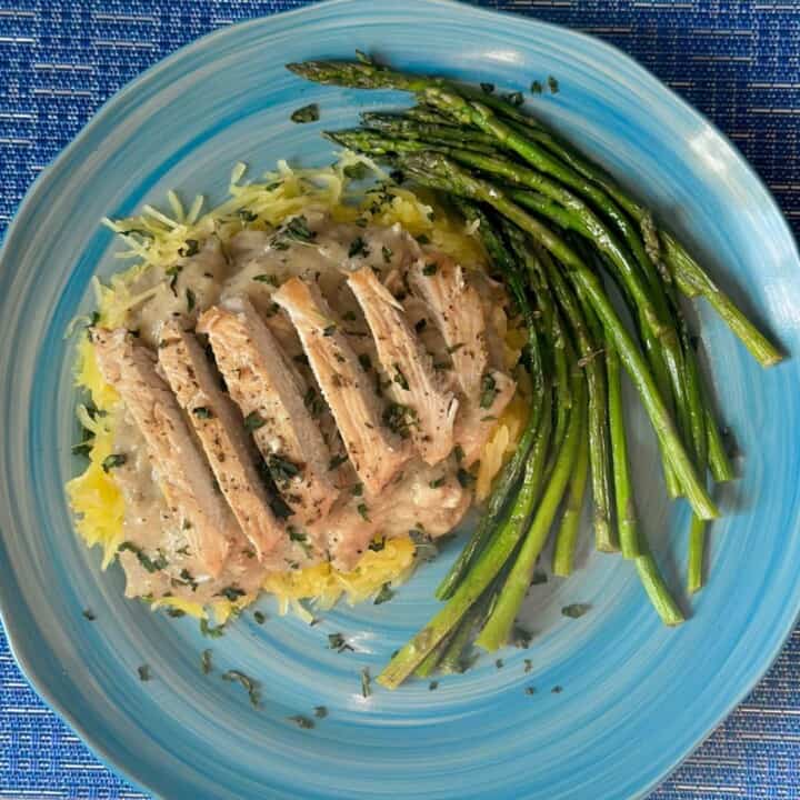A closeup photo of chicken alfredo on a teal plate. Cooked spaghetti squash is topped with a creamy tan alfredo sauce mixed with chunks of chicken breast. On top is a line of sliced chicken breast garnished with freshly chopped parsley. Next to the main dish is a swirl of cooked asparagus drizzled in avocado oil and seasoned with salt and pepper.