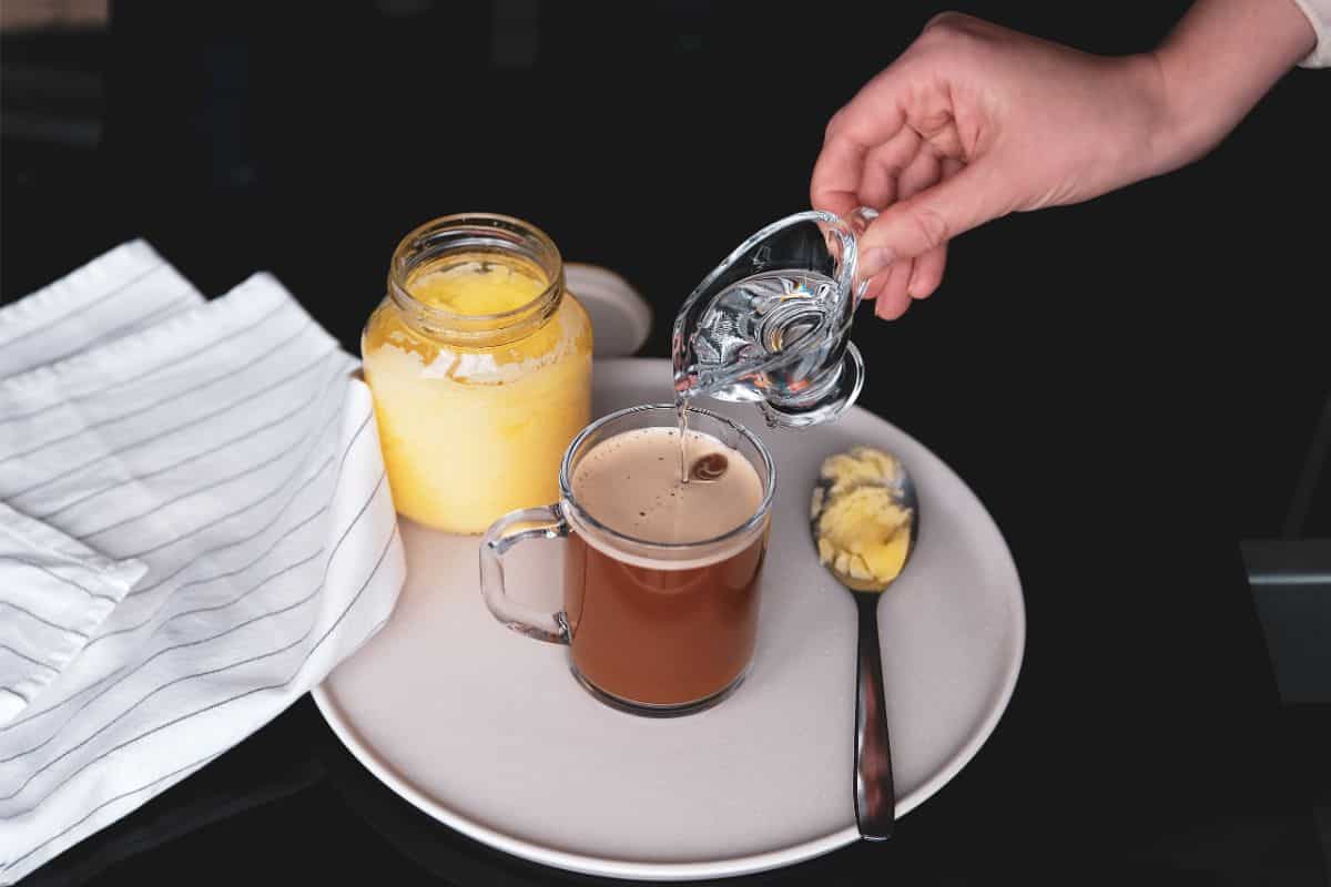 A white plate has a clear glass coffee cup with coffee in it. A woman's hand is pouring a clear MCT liquid into the coffee. Next to the coffee is a glass jar of ghee and a spoonful of ghee to the right. To the left of the plate is a white and black striped kitchen towel all on a black countertop.