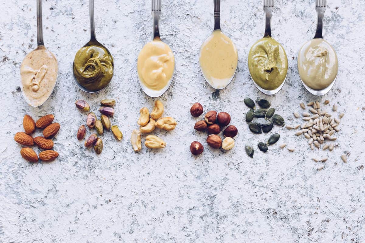 A photo of 6 spoons full of 6 different nut butters and in front of them are a small handful of the seed that created the butter. The seeds and butters are in various colors and consistencies.