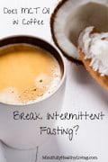 A pinterest optimized photo of A white coffee mug with frothy coffee in it ontop of a white wood table next to a wooden spoon with MCT containing coconut oil in it. Next to the spoon is half of a coconut. Text overlay says Does MCT oil in coffee break intermittent fasting? mindfullyhealthyliving.com