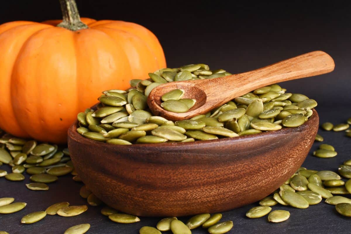 A bowl of shelled pumpkin seeds with a wooden spoon in it. The seeds are spilling over the table. Next to the bowl is an orange pumpkin.