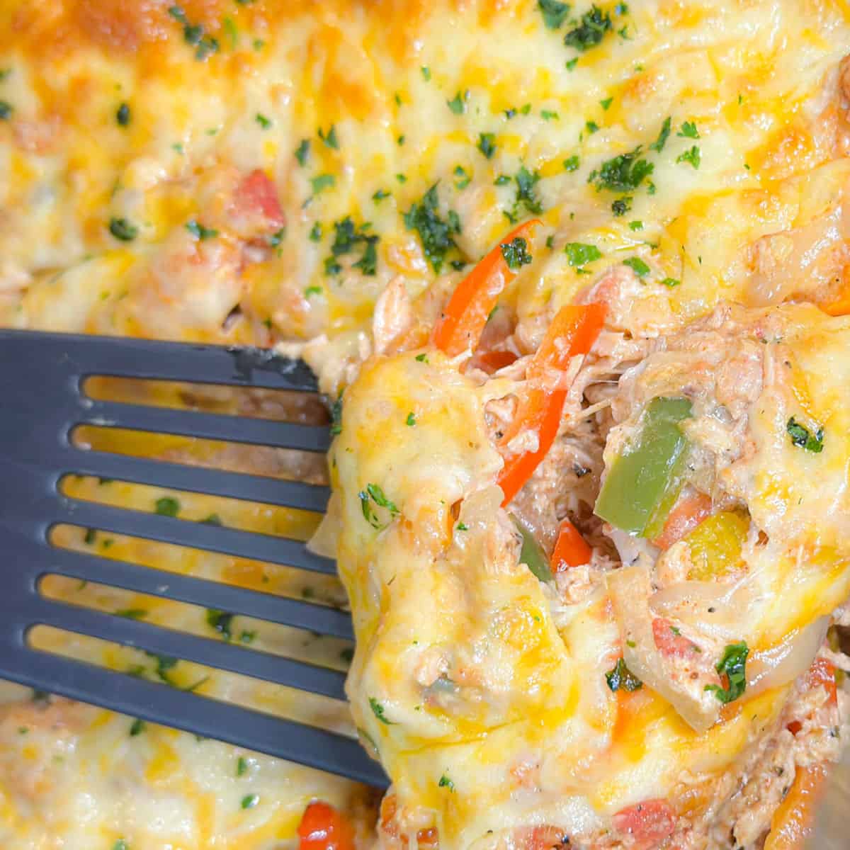 A decadent closeup of chicken fajita casserole melted with cheese being scooped by a black spatula. Gooey cheese covers shredded chicken mixed with colorful peppers and creamy sauce garnished with cilantro