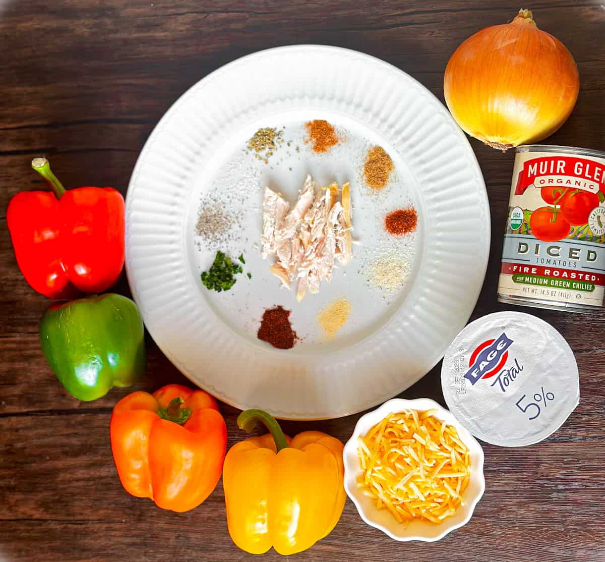 Photo of ingredients for chicken fajita casserole with a green, yellow, orange, and red bell pepper, a ramekin of shredded cheese, a container of Fage 5% yogurt, a can of diced tomatoes fire roasted with medium green chiles and a yellow onion surrounding a white plate with shredded chicken in the middle and various spices that make up the homemade fajita seasoning for this recipe.