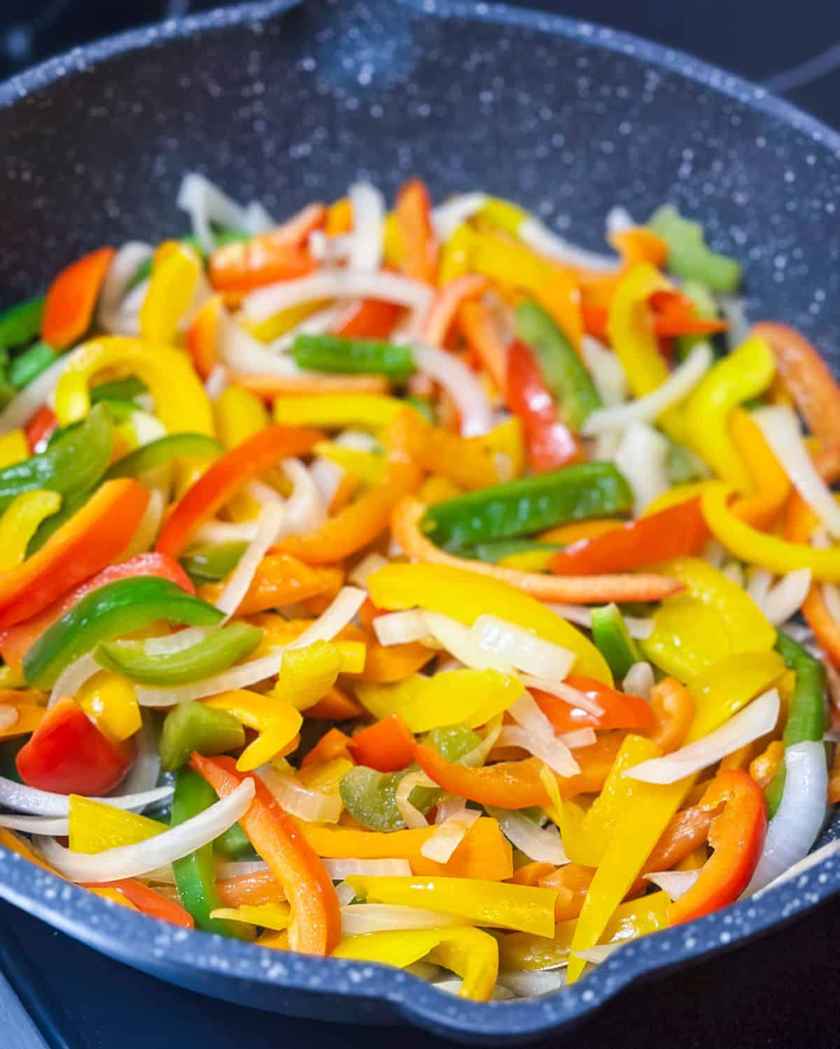 slices of colorful peppers and white onions are sauteeing in a speckled grey skillet on a black top stove.