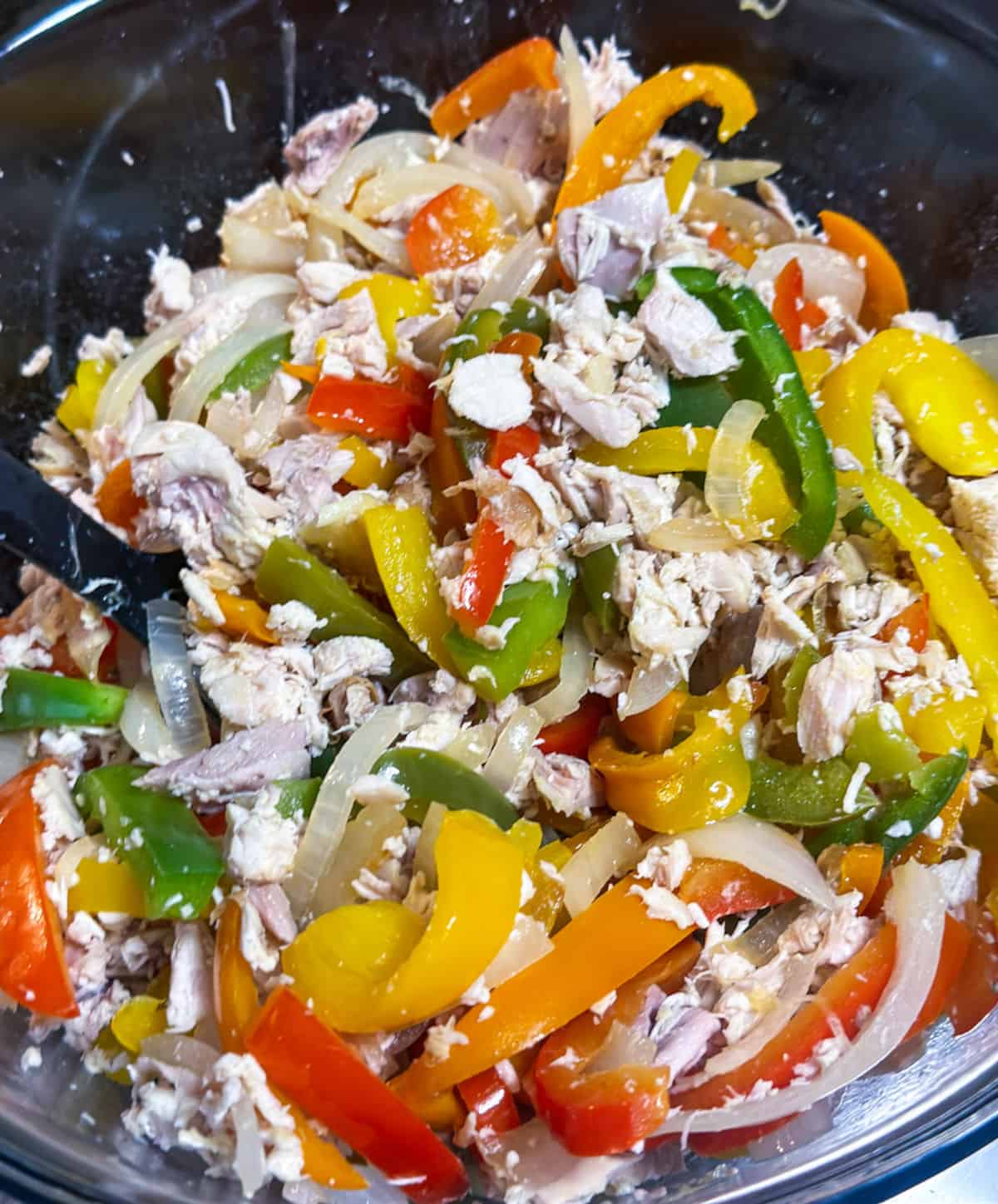 Shredded chicken is mixed with sauteed onions and bell peppers of all colors in a glass bowl for the second step in the recipe.