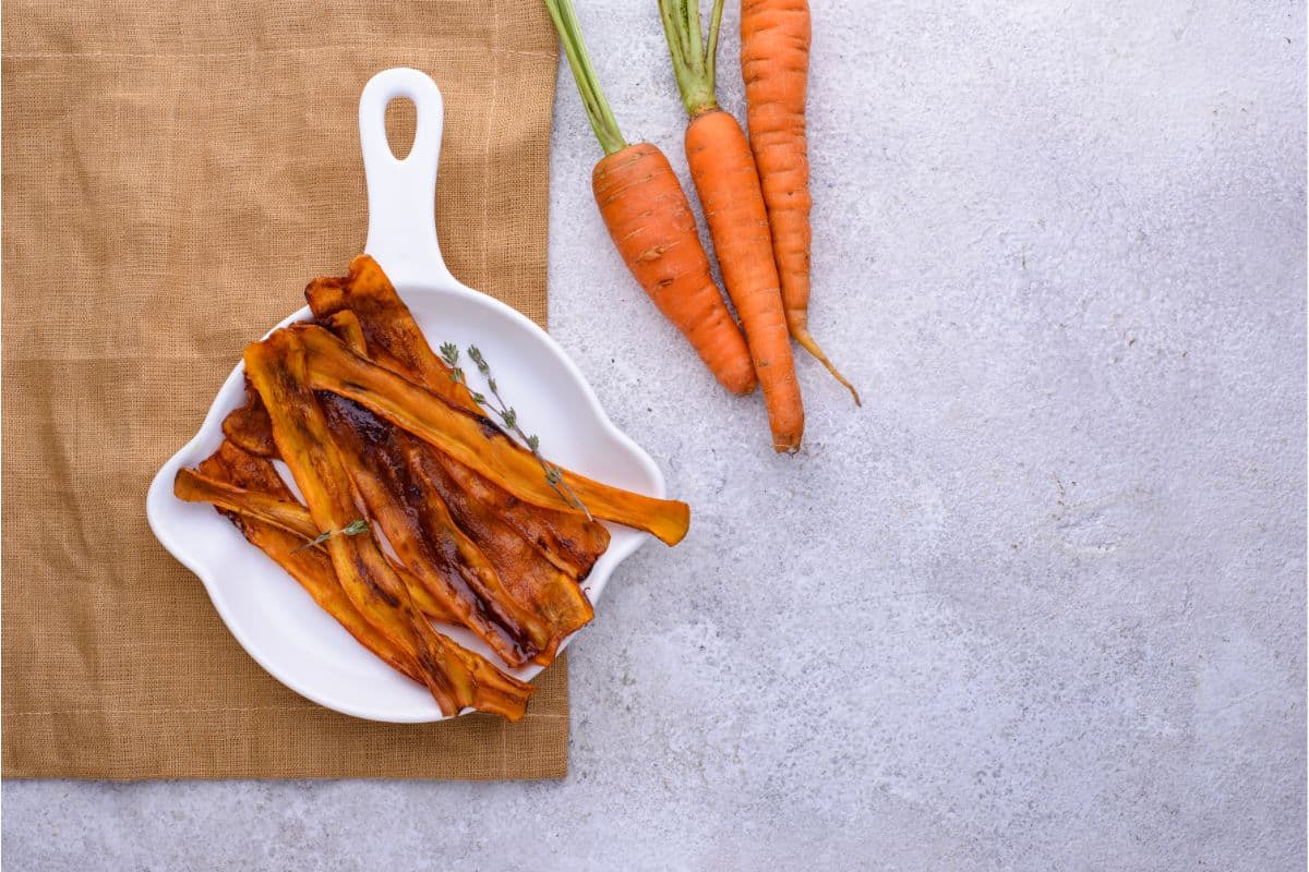 This photo is an example of plant-based bacon made from sliced carrot and cooked with sauces that make them look and taste like the flavors of bacon. The strips are on a white plate next to a group of 3 raw carrots