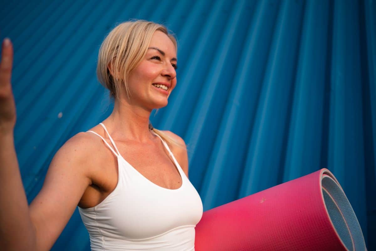 A smiling woman in a white tank top with a yoga mat depicting lowered stress levels and balanced hormones.