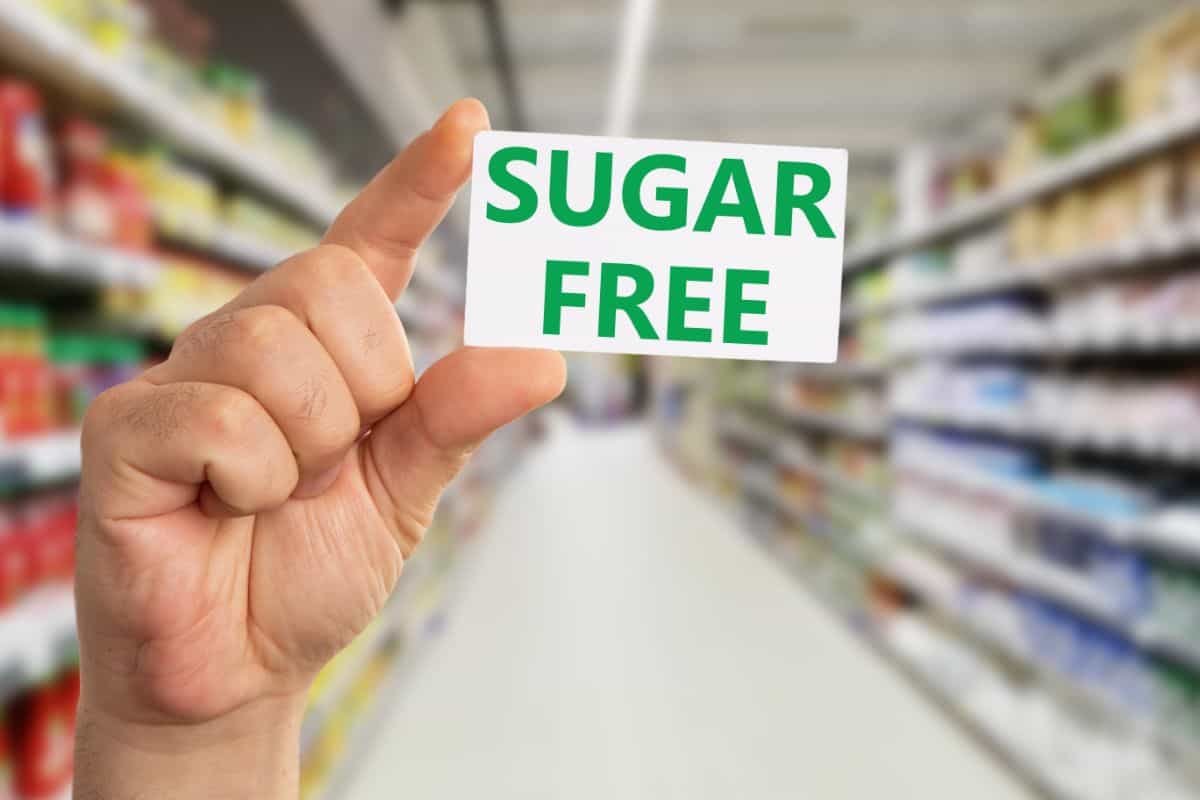 A close up of a male hand holding a white card with his thumb and forefinger that reads in all green caps SUGAR FREE. In the blurred background appears to be grocery store shelves.