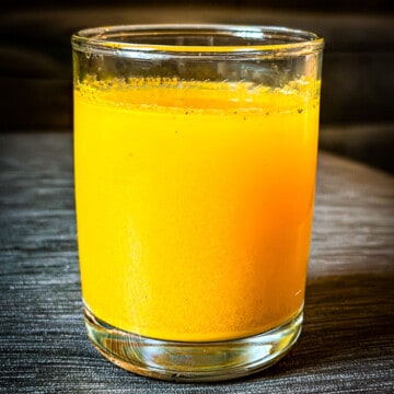 A clear glass cup with brightly colored yellow and orange turmeric lemon juice with a few specs of visible black pepper added for absorption.