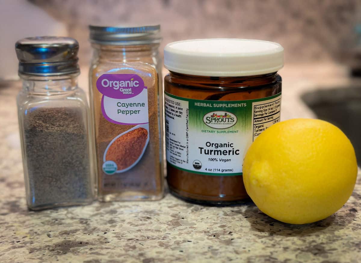 An ingredients photo of a clear pepper shaker with black pepper in it next to an organic cayenne pepper shaker, a rounder short jar of organic turmeric powder and one whole yellow lemon.