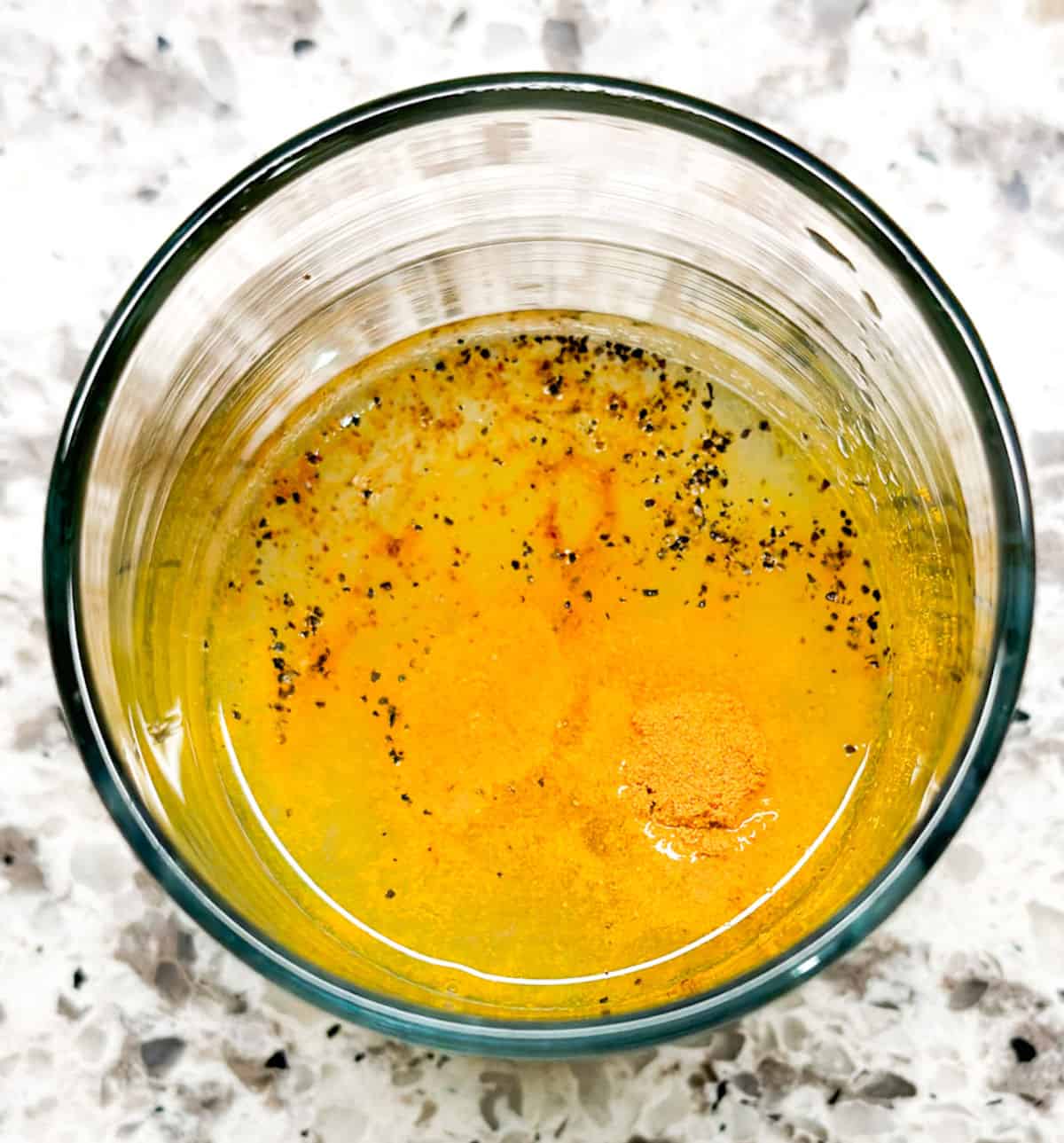 A birds eye view of a clear glass with lemon juice in it. On top of the juice is a scoop of turmeric powder, a splash of cayenne pepper, and a pinch of black pepper sprinkled in it before it is stirred.
