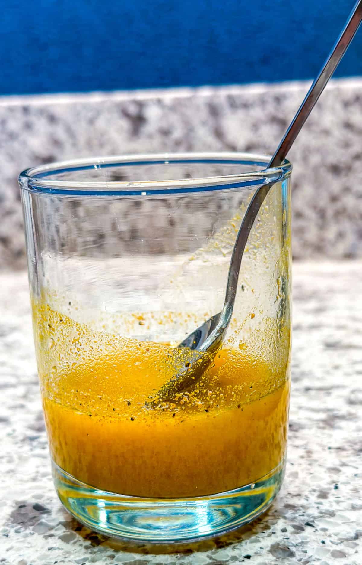 A glass cup with a spoon in it that is one quarter full of orange colored turmeric lemon juice with pepper and turmeric powder on the sides from stirring.