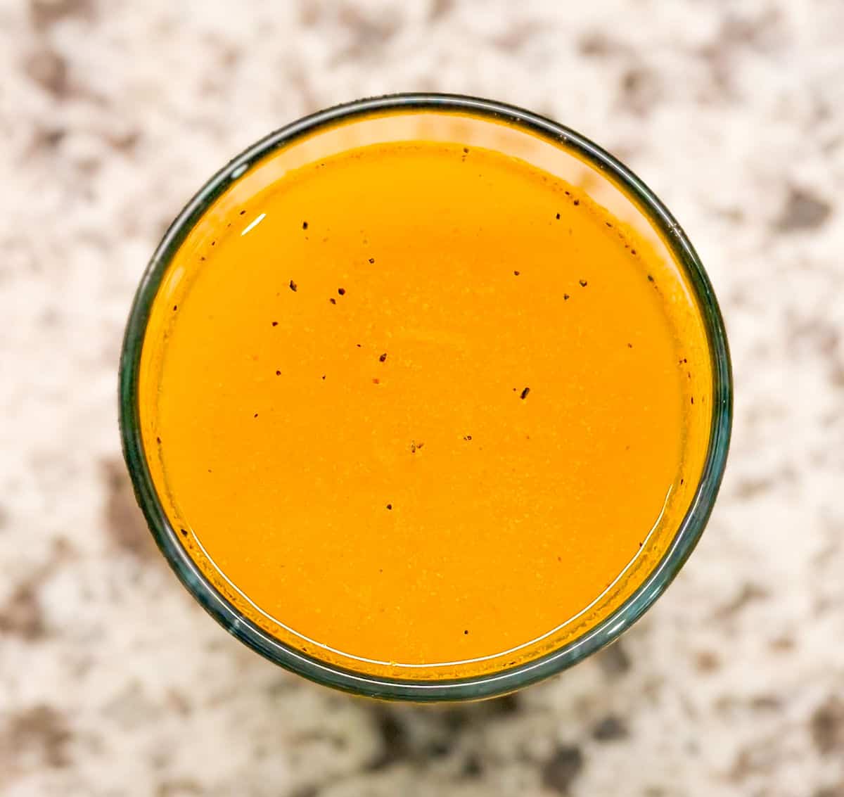 a birdseye view of the glass of turmeric lemon juice after adding water to the mixture. a few specs of black pepper float in the orange drink.