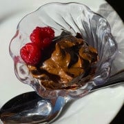 Chocolate Avocado Pudding with a spoon and white plate and two red raspberries on top in a clear decorative bowl with chocolate chips on the side