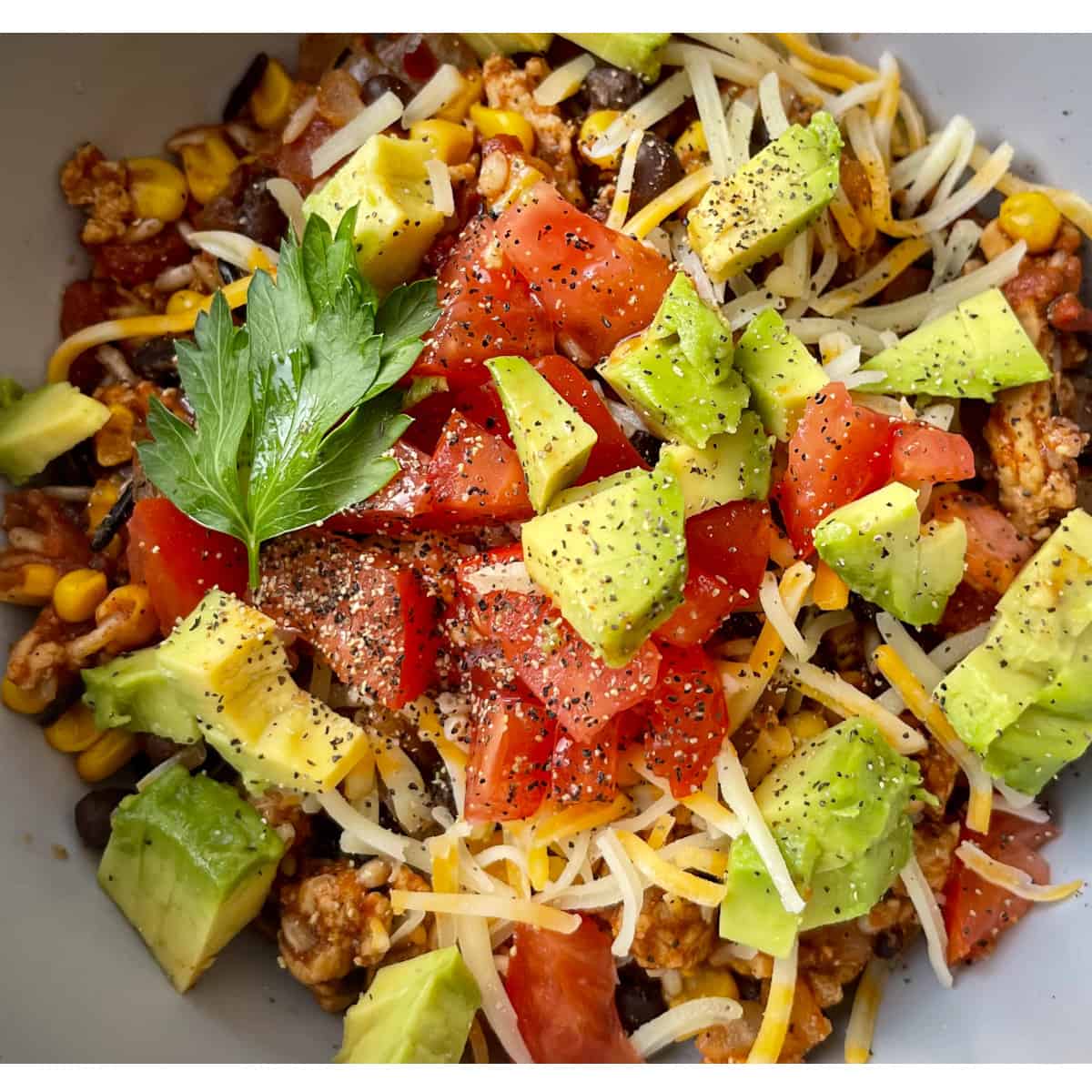 A prepared bowl of Healthy Cheesy Taco Skillet up close garnished with black pepper, tomato, avocado, cheese and a cilantro leaf