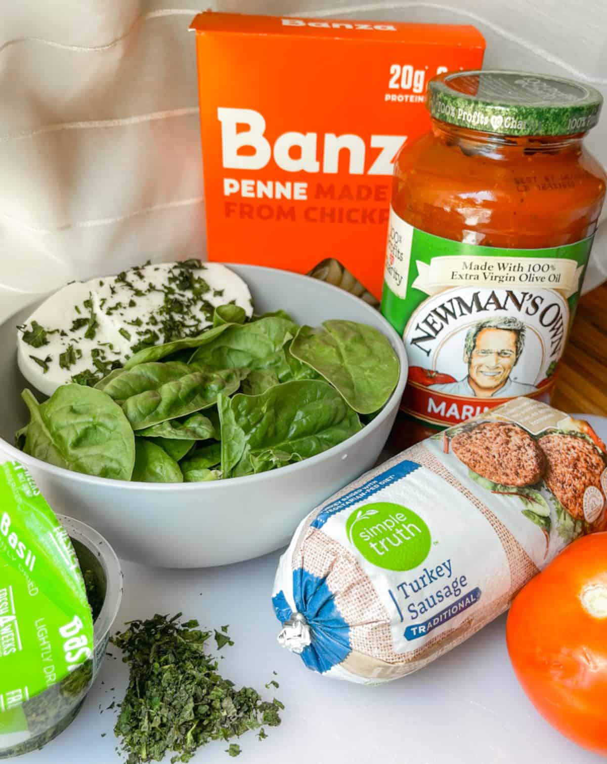 fresh spinach, and fresh mozzarella in a bowl, box of Banza penne, Newmans own marinara sauce, simple truth traditional turkey sausage, and basil Blakes lightly dried on a cutting board- all ingredients ready for healthy spinach and sausage skillet