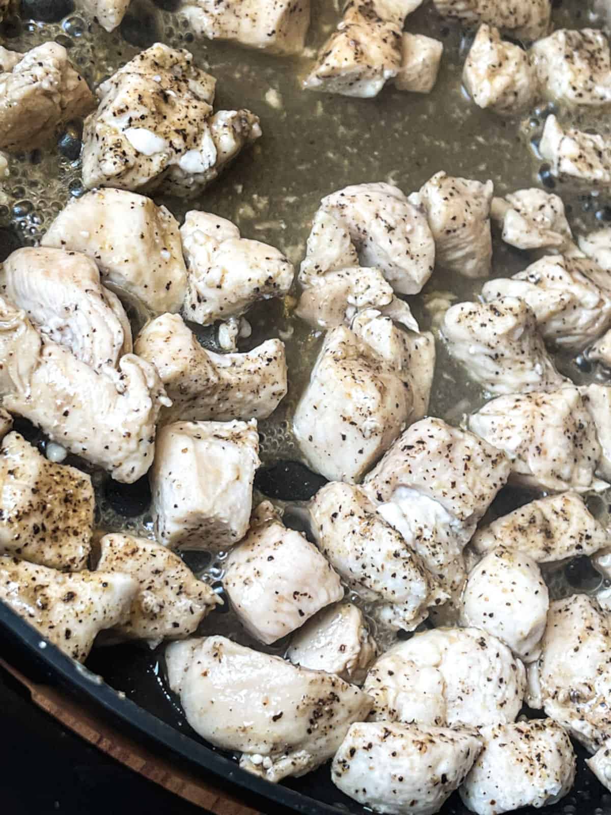chicken breast cut into cubes with black pepper cooking in a skillet with liquid seen in the pan