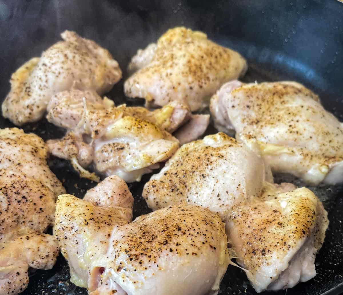 6-7 chicken thighs seasoned with black pepper cooking in a black pan greased with oil. steam can be pictured, indicating it is actively cooking in a 2nd step prep for Creamy Tuscan Chicken Recipe