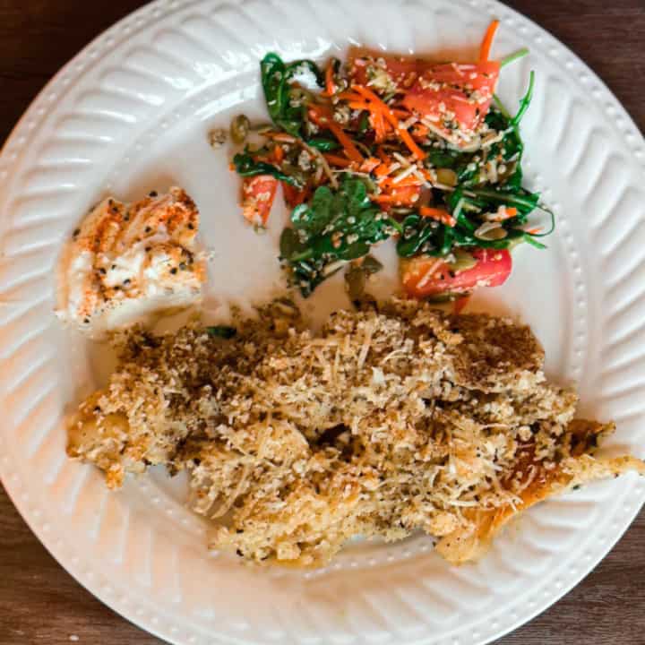 A decorative round white plate on a wooden table with parmesan crust topping over fish at the bottom, lemon cayenne yogurt sauce at the top left and a green arugula side salad with cut tomatoes, shredded carrots, hemp seeds, and pumpkin seeds with a vinegarette dressing for final product of Keto Parmesan Pollock