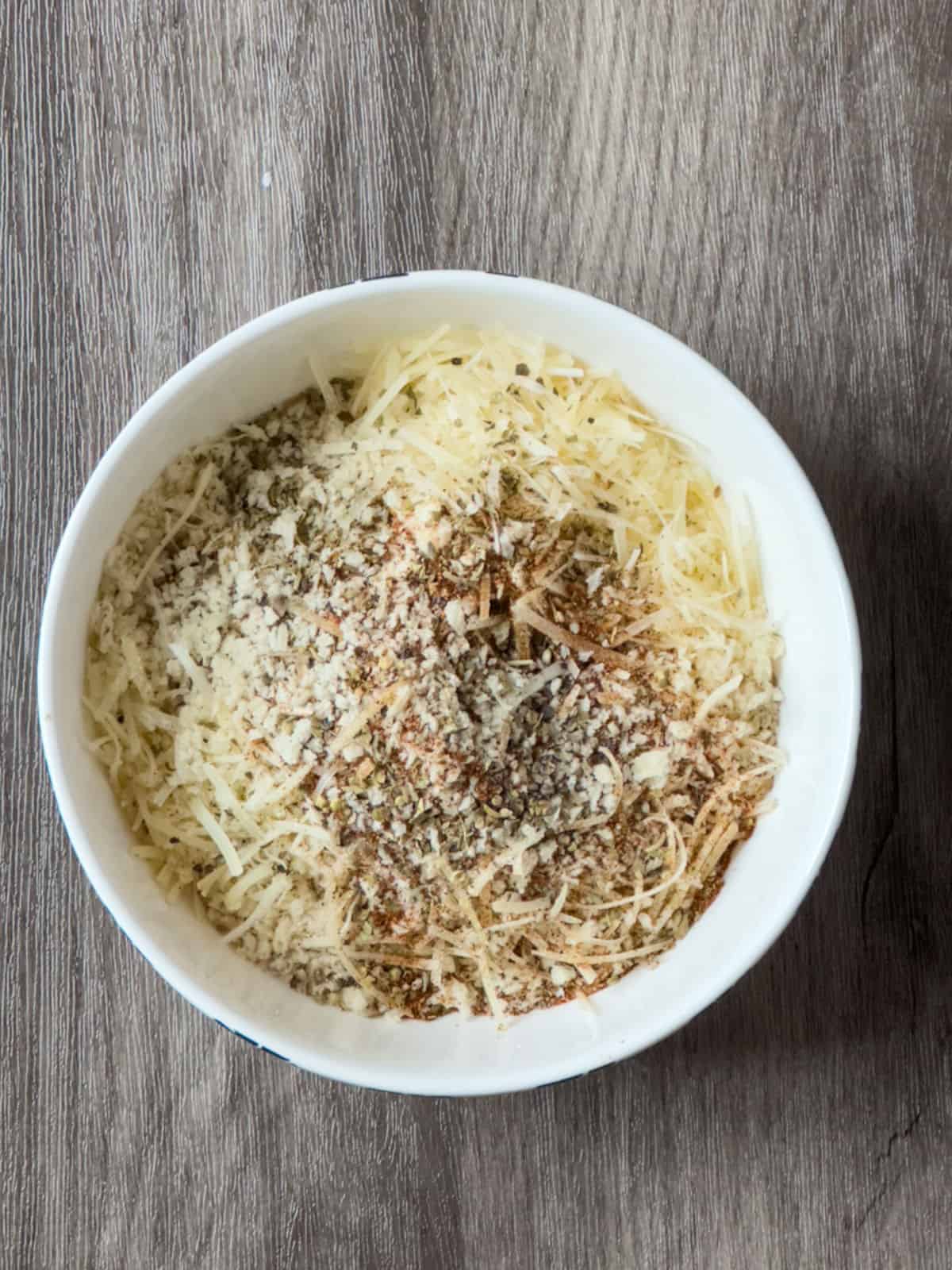 a white bowl on a wooden background with seasoning, shredded parmesan and panic bread crumbs mixed together