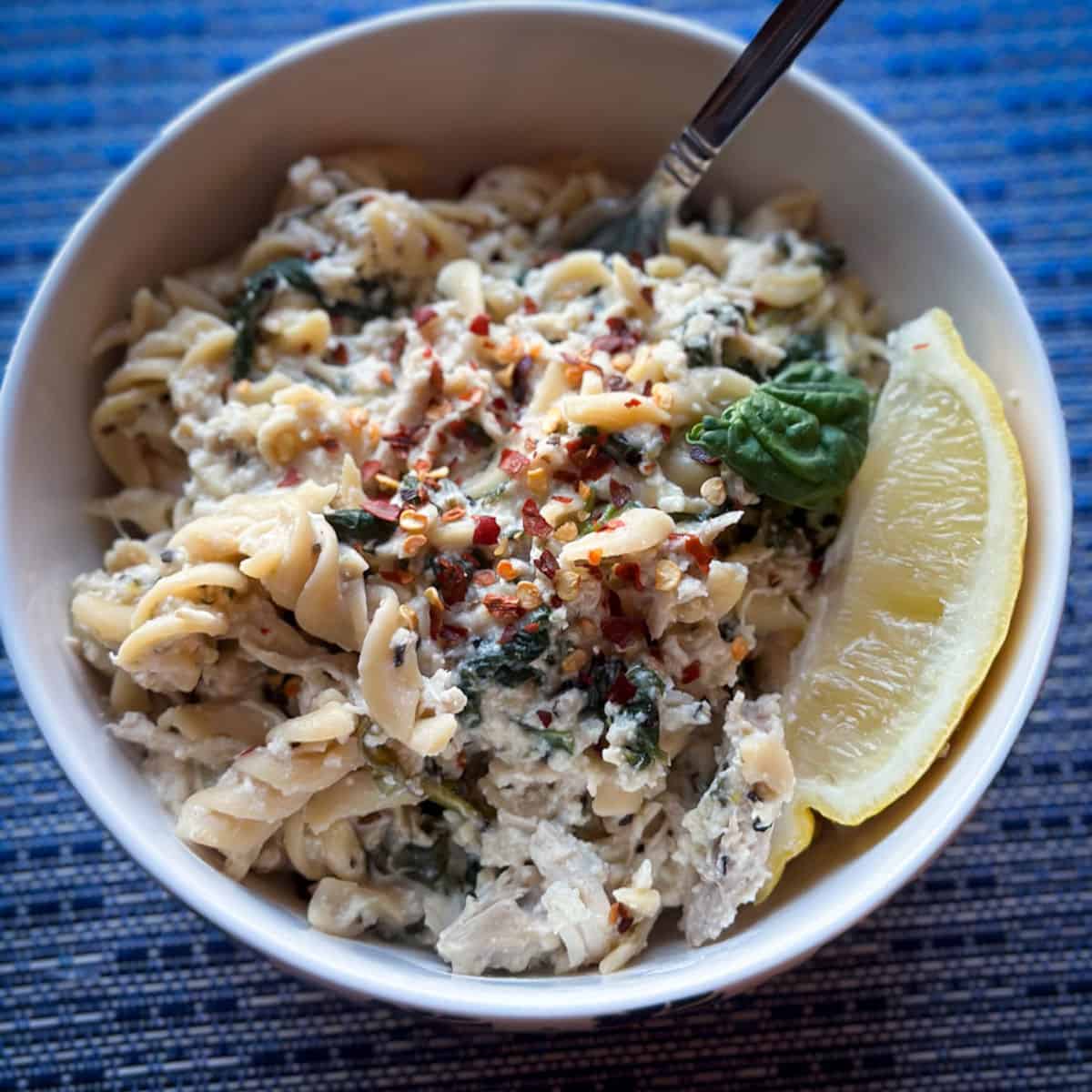 a bright blue placemat with a white bowl and creamy lemon ricotta pasta with chicken spinach ad red pepper flakes with. lemon wedge and fork in the bowl
