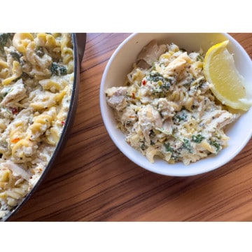 a pot filled with creamy lemon ricotta pasta in the left corner next to a white bowl of the pasta with a lemon wedge in it all on a wooden table