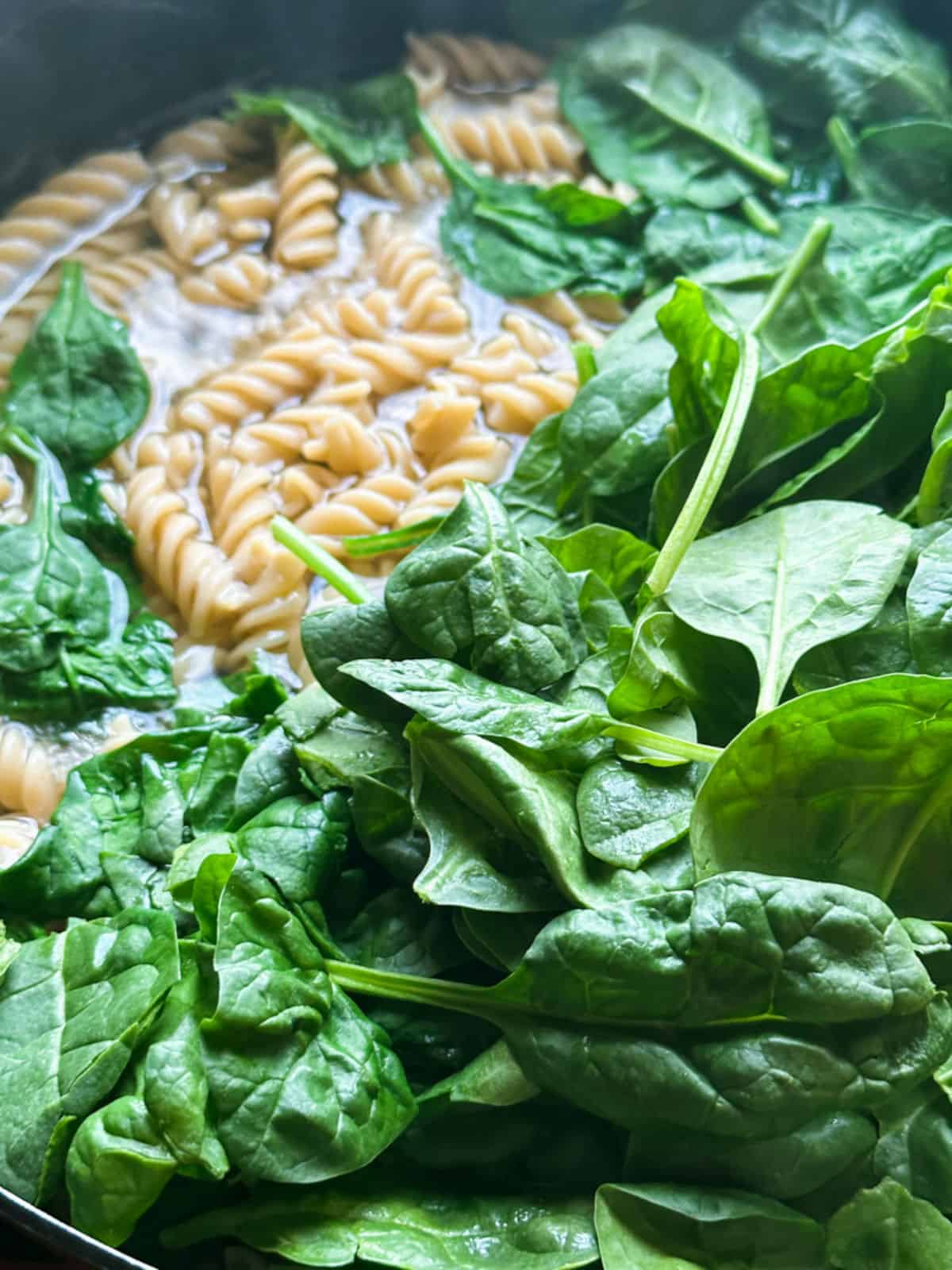 a pot with rotini pasta cooking in water with fresh spinach added