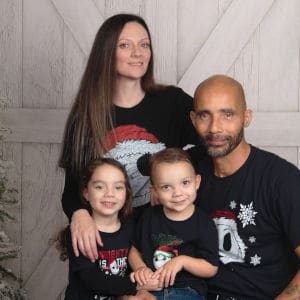 a woman standing and a man sitting with two toddlers in front in a family photo. all wearing black shirts with nightmare before Christmas character jack skellington on the front. with a light wood backdrop