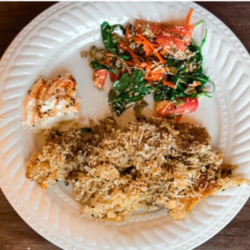 A decorative round white plate on a wooden table with parmesan crust topping over fish at the bottom, lemon cayenne yogurt sauce at the top left and a green arugula side salad with cut tomatoes, shredded carrots, hemp seeds, and pumpkin seeds with a vinegarette dressing