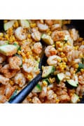 Spicy cajun shrimp with zucchini, corn cooking in a skillet stirred by a black spoon