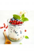 an open jar of yogurt with granola at the bottom mixed with red and blue berries topped with red and blue berries on a twig with a mint leaf for garnish. a wooden spoon and red berries in the background can be seen