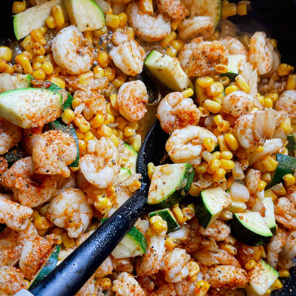a skillet cooking shrimp, zucchini and corn seasoned with cajun spices with a black cooking spoon taking a spoonful