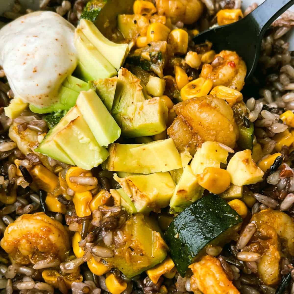 spicy cajun shrimp bowl with cooked shrimp, corn, green zucchini on rice with chopped avocado and a dollop of yogurt sauce with red specs in the top left corner a spoon sits in the rice in the top right corner