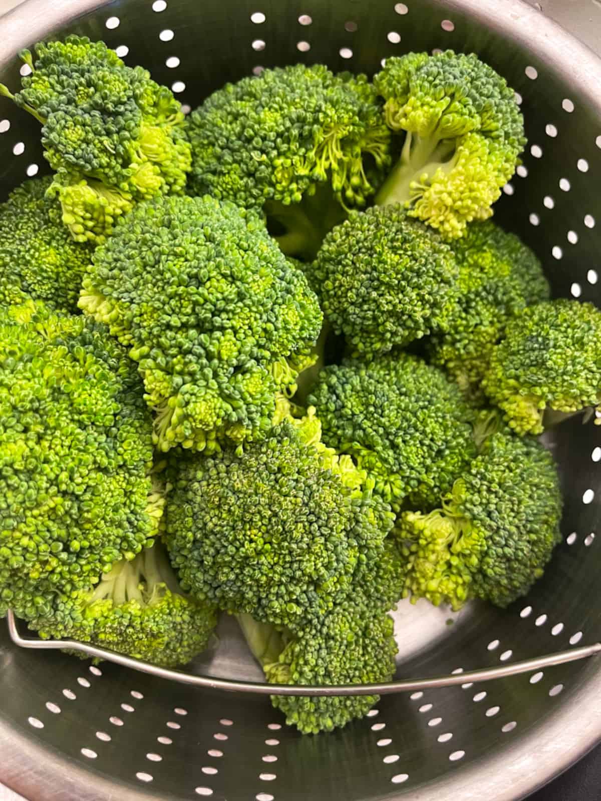 raw broccoli florets in a metal steamer basket with holes and a metal handle