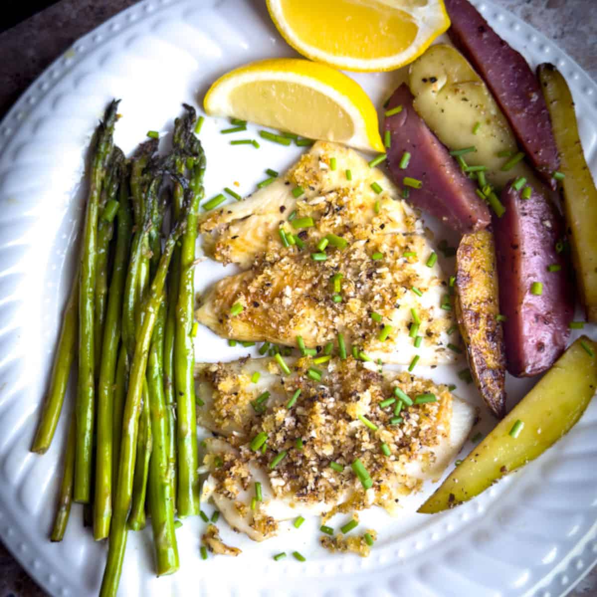 a white plate with a marble counter with yellowtail flounder with herbal crush and chives on top with two slices of lemon and a handful of slice fingering potatoes, cooked and seasoned on the right