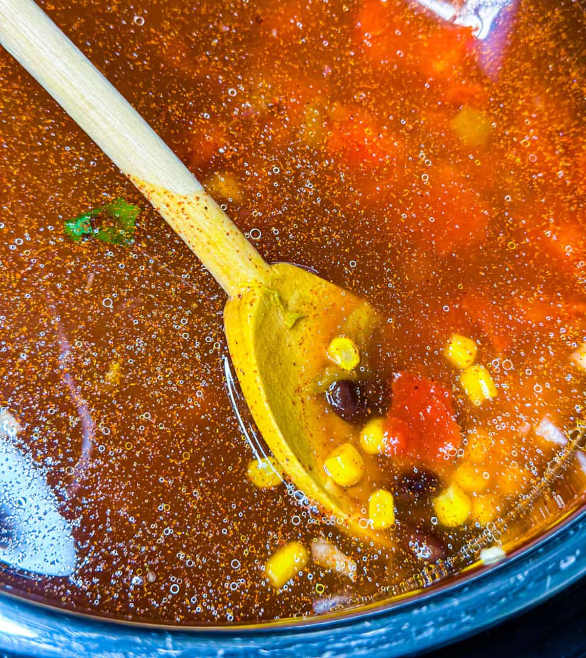 a silver pot with red and orange liquid soup with a wooden spoon scooping tomato, corn, and black beans with spices throughout the pot