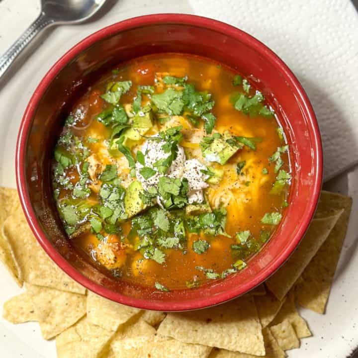 a deep red bowl with soup with chunks of chicken, melted cheese, cilantro, chopped avocado, sour cream with pepper on it. under the bowl is a handful of white corn tortilla chips with a silver soon partially visible at the top left corner and a white napkin at the top right corner