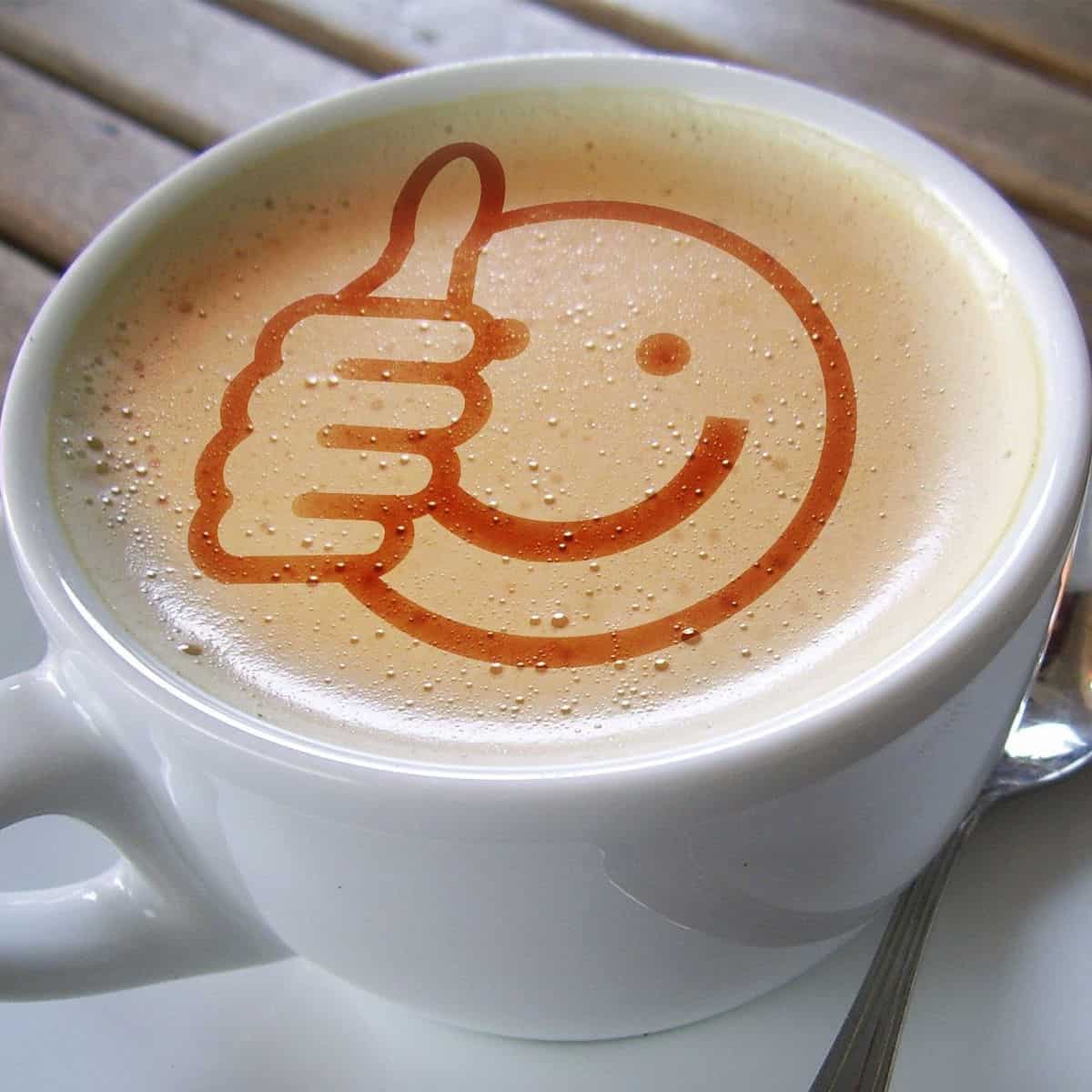 A white coffee mug with a silver spoon next to it on a wooden table backdrop. there is tan foam and a dark tan smiley face drawing with a thumbs up to the left of the face in thefoam
