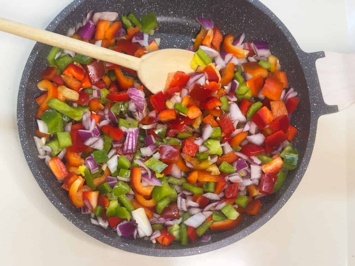 A grey with white speckled saute pan with a light tan handle pointing left with chopped red onions and green and red bell peppers ready to saute on top of melted ghee with a light wooden spoon in it