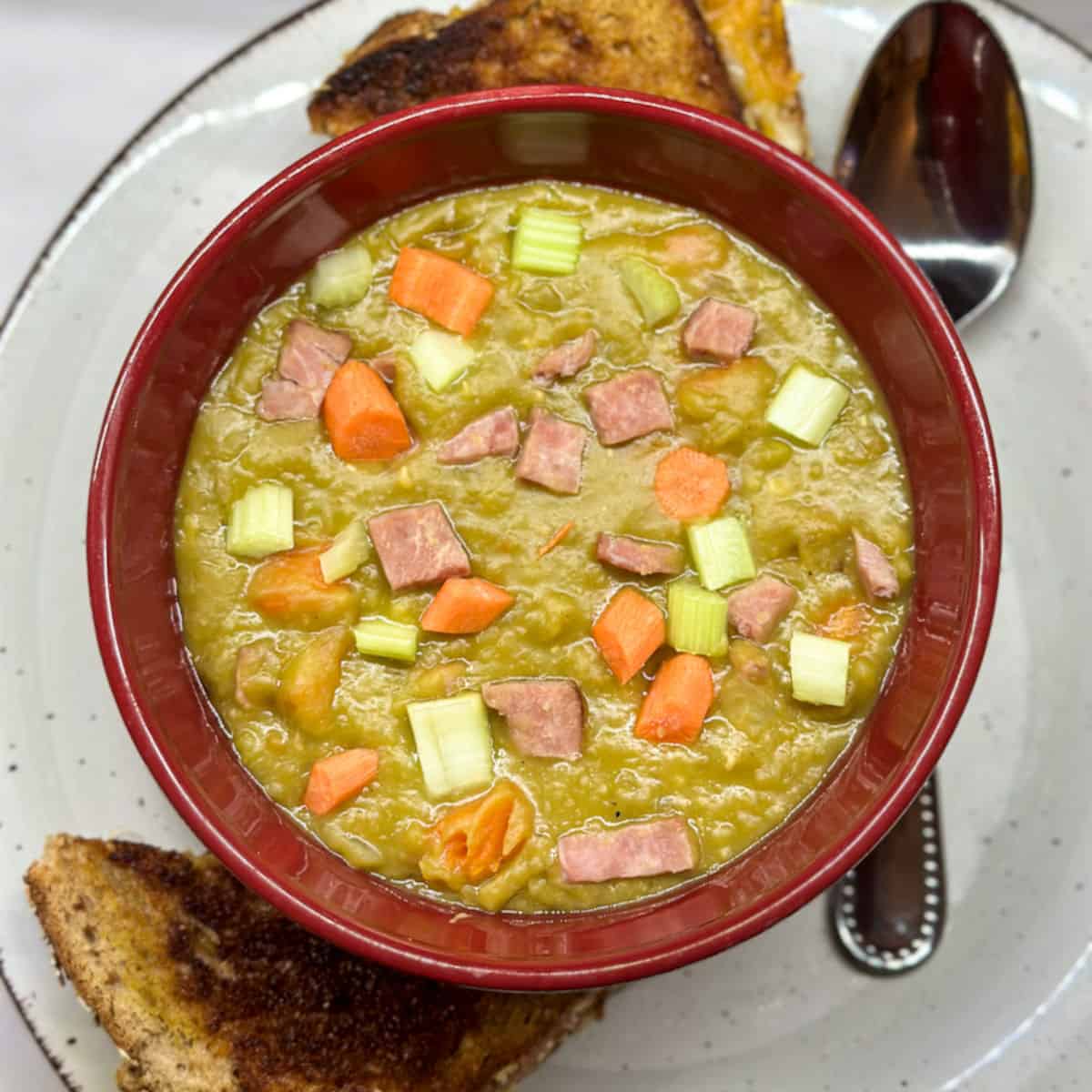a red bowl on an of white plate with brown outline and a corner cut grilled cheese sandwich halves at the top and bottom of the plate and a silver spoon on the right. the bowl is filled with a thick pea green soup with ham, carrot and celery chunks sprinkled on top