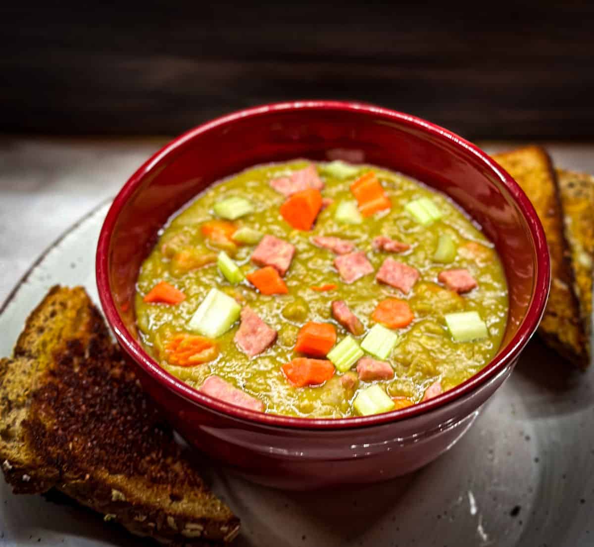 a red bowl on an of white plate with brown outline and a corner cut grilled cheese sandwich halves at the top and bottom of the plate and a silver spoon on the right. the bowl is filled with a thick pea green soup with ham, carrot and celery chunks sprinkled on top