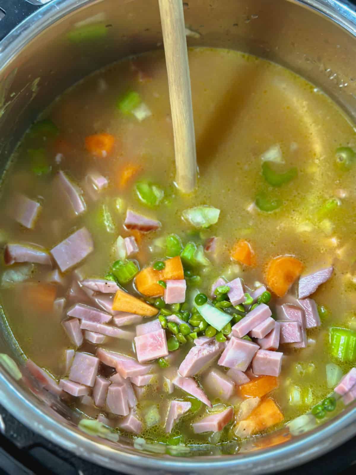 a silver instant pot bowl with a light wooden spoon immersed in the light colored amber soup broth with chopped ham, uncooked split peas, carrots, and celery