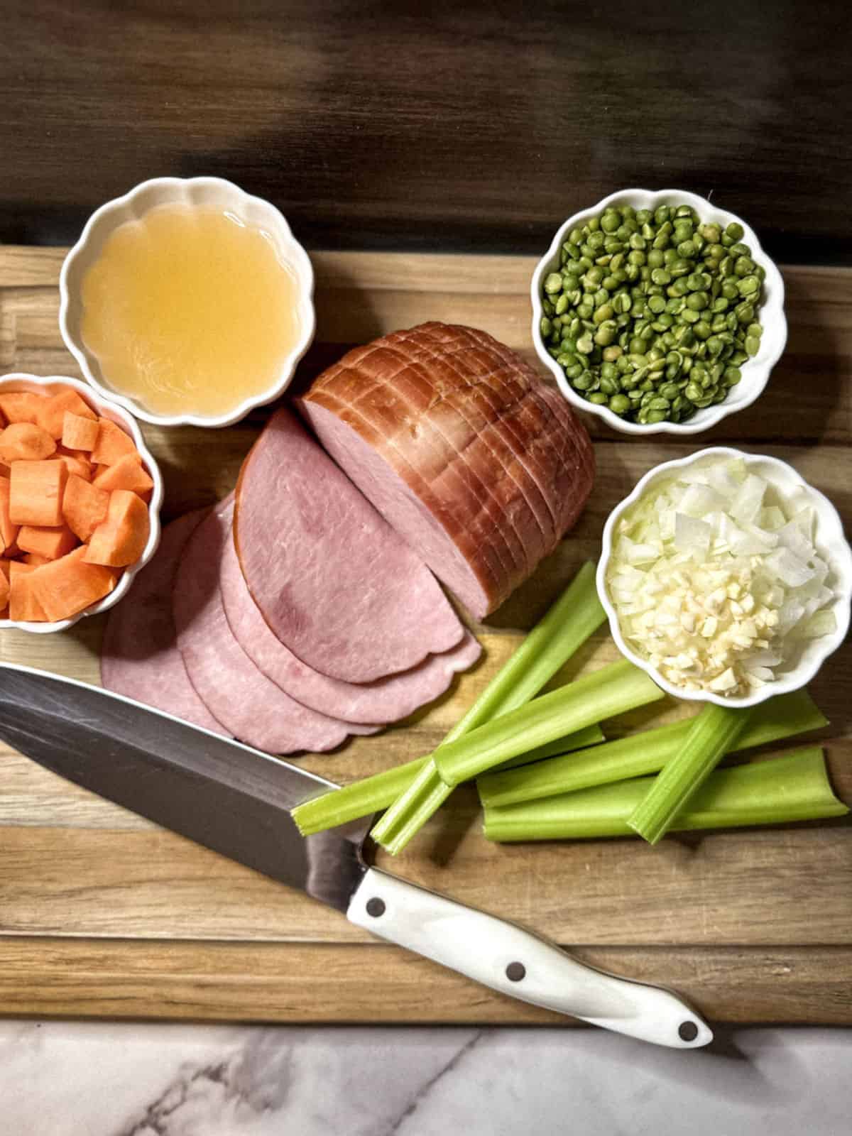 the ingredients for split pea soup on a light wood colored cutting board with a dark wood colored backsplash and a white marble counter and a white handled cutco chef knife at the bottom with sliced half ham in the middle, and a few cut celery stalks off to the right with 4 white ramekins with (left to right) chopped carrots, broth, rinsed green split peas, and chopped onions
