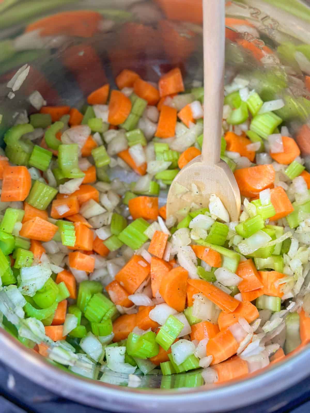 a silver instant pot with a light wooden spoon with almost raw chopped celery carrot and onion likely satueing in ghee