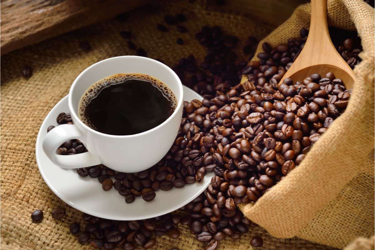 a burlap placemat with a white coffee cup and saucer with dark coffee in the cup and brown coffee beans spilling all around the saucer and burlap sack