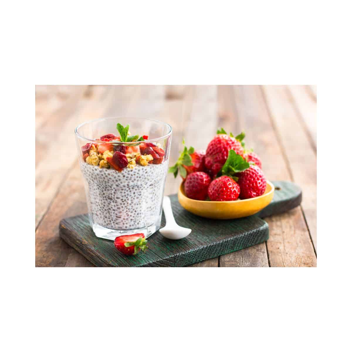 a bright photo of a clear glass with chia seeds hydrated in milk with various fruits on top of the chia pudding. next to the cup is a white spoon and a yellow dish with a pile of strawberries on it. all of this is on a rustic green board atop a wooden table