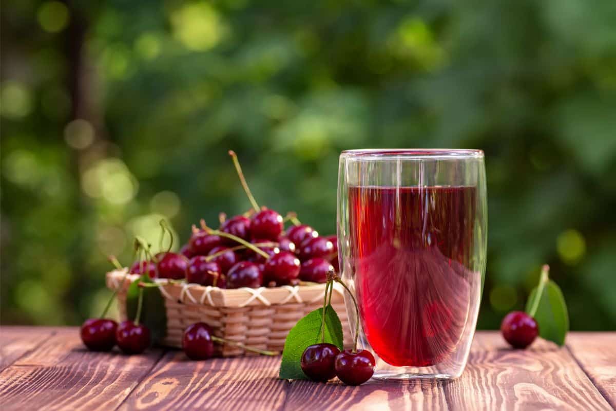 An outside wooden picnic table with lots of greenery in the background. On the table is a light colored wicker basket with cherries with stems on them in and around it and in front of the basket is a clear glass full of red cherry juice