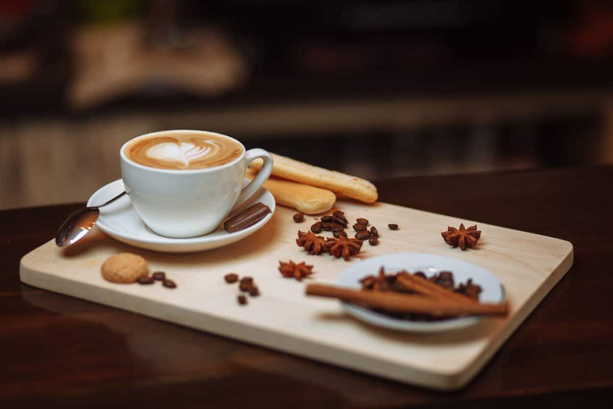 A dark cherry wood table with a small light wood board with a small white coffee cup and saucer with a silver spoon on the left with coffee and a decorative white creamer heart in the cup and a piece a chocolate on the saucer. two oblong cookie biscuits behind the cup and one small round one in front of it. to the right are small piles of a few coffee beans and decorative flowers and a white blurred saucer in the front right with 3 cinnamon sticks on it
